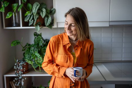 Pleased woman enjoy morning routine with coffee at home rest in kitchen surround by houseplants looking aside. Satisfied joyful middle aged female standing at home have pleasure in calmness cozy home 