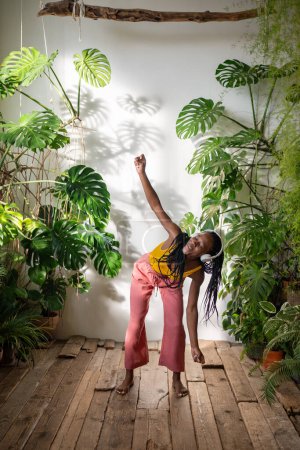 Photo for Carefree black woman dancing barefoot in headphones listening to music on wooden floor with pleasure, enjoy moment. Relaxed african american young female chilling out to songs in room with houseplants - Royalty Free Image
