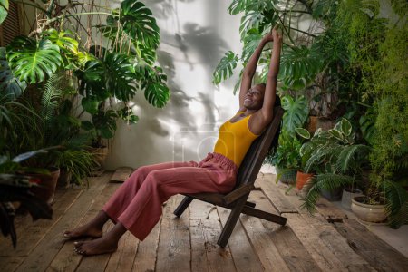 Satisfait femme afro-américaine étirant les bras vers le haut reposer sur la chaise entourée de plantes tropicales d'intérieur. Femme noire détendue s'assoit dans la chambre avec l'intérieur de la jungle urbaine profiter du temps tranquille avec des plantes d'intérieur.