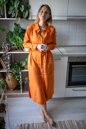 Photo for Satisfied woman plants lover rest in kitchen holding cup of coffee looking at camera. Relaxed pleased scandinavian middle aged female standing at home enjoy calmness morning surround by houseplants. - Royalty Free Image