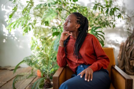 Pensive african american woman concentrated sitting on armchair taking break, planning thinking surround by houseplants at home. Thoughtful relaxed black girl rest in room with urban jungle interior.