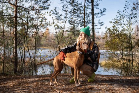 Photo for Middle aged woman petting Hungarian Vizsla dog walking in nature autumn park. Female travelling spending time with pet friend on weekend sitting on haunches. Travel, tourism with dog, wanderlust. - Royalty Free Image