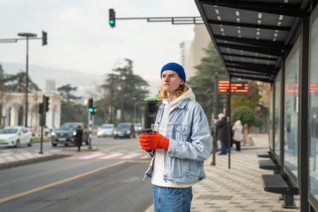 Téléchargez les photos : Un hipster aux cheveux longs en tenue élégante, debout dans un arrêt de bus avec une tasse de café, regardant au loin, attendant un taxi, un bus. Homme à la mode insouciant, représentatif de la génération z - en image libre de droit