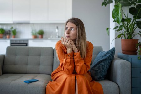 Anxiety woman in stress, looking aside nervously squeezes hands sitting on sofa thinking about problems after receive bad news on cellphone. Worried female feel frustration waiting call on smartphone.