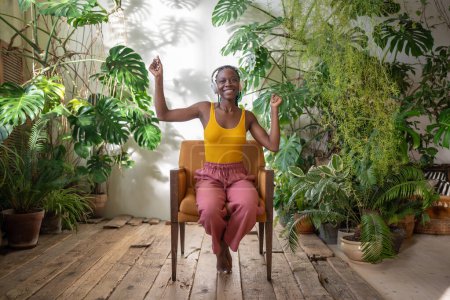 Unbekümmert fröhliche Afroamerikanerin mit Kopfhörern, die Musik inmitten von Zimmerpflanzen genießt. Fröhlich lächelndes schwarzes Mädchen sitzt auf Stuhl zu Hause tropischen Garten hören Lieblingslied und leicht tanzen.
