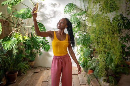 Joyeuse blogueuse afro-américaine prend selfies entourer cultivées plantes tropicales d'intérieur pour blog. heureux noir fille plantes amant sourire faire auto photo sur téléphone mobile dans la maison jardin jungle urbaine.