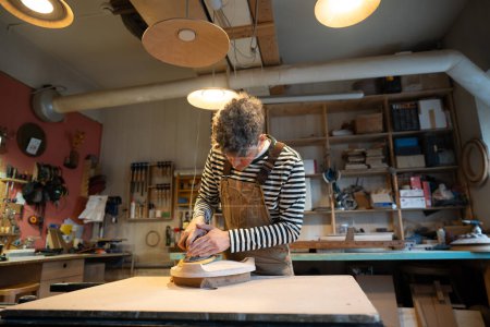 Holzschleifen mit Orbitalschleifer in der Werkstatt. Konzentrierte Mann Schreiner poliert Holzsitz eines zukünftigen Stuhls mit Elektroschleifer. Tischlerei. Möbelproduktion