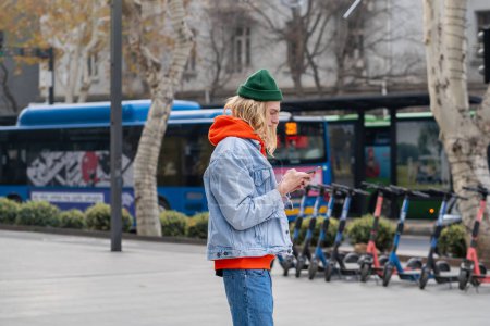 Photo for Trendy long-haired hipster guy in colorful stylish hoodie standing in city street, holding smartphone, reading, messaging, scrolling web pages, photos in internet, spending time in social networks - Royalty Free Image