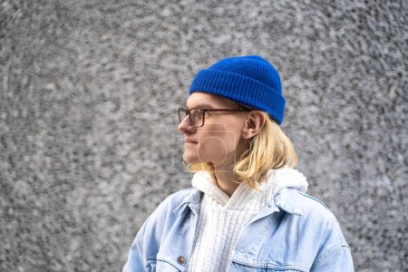 Téléchargez les photos : Homme hipster blond aux cheveux longs avec des lunettes élégantes debout dans la rue de la ville au-dessus du mur gris, regardant au loin. Portrait rapproché de mâle à la mode ressemble modèle, étudiant international, touriste - en image libre de droit