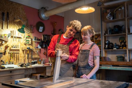 Interested carpenter couple discusses working moment with smile. Attentive male joiner looks at work product of female woodworking apprentice, gives instructions, advises. Teamwork, small business.