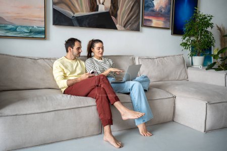 Photo for Pleasant family couple artists sits on couch looking at laptop screen browsing internet together. Creative young married spouse web surfing, making purchases online, booking flight tickets for travel. - Royalty Free Image