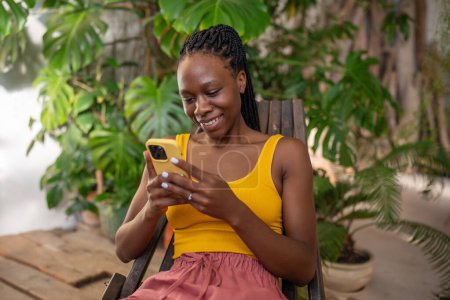 Téléchargez les photos : Cheerful relaxed African American girl rest takes break with smartphone among grows indoor plants spending time online on Internet. Happy Black woman smiling using cellphone text social media message. - en image libre de droit