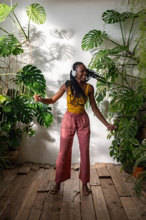 Unbekümmertes schwarzes Mädchen, das barfuß in Kopfhörern tanzt und mit Vergnügen Musik auf dem Holzboden hört, genießt den Moment im Gewächshaus. Entspannte Afroamerikanerin chillt zu Hause mit Pflanzen zum Singen