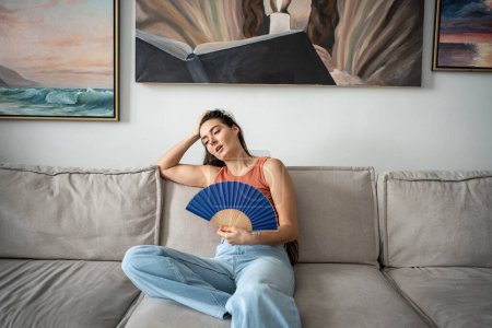 Overheated woman sits on couch in living room at hot summer weather day feel discomfort suffers from heat waving blue fan to cool. Exhausted girl artist sweating cannot working without air conditioner