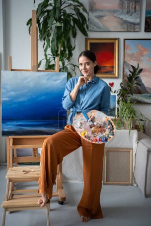 Zufriedene Künstlerin lächelnd in Kamera mit Farbpalette steht neben Staffelei mit Leinwandbild im Kunstatelier. Fröhliche Malerin mit Pinsel in Bastelwerkstatt lenken Prozess ab.