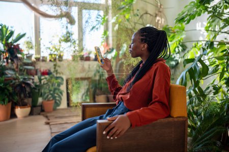 Téléchargez les photos : Satisfait femme afro-américaine détendue à l'aide d'un smartphone assis sur un fauteuil confortable entouré de plantes d'intérieur. Agréable repos féminin noir avec téléphone mobile surfer sur les médias sociaux dans le jardin de la maison confortable. - en image libre de droit