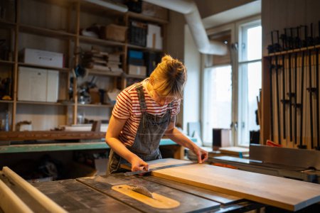 Téléchargez les photos : Femme focalisée charpentier attentif prépare la planche en bois à travailler sur des paramètres de mesure de scie circulaire avec règle. Menuisier pour couper le bois sur l'équipement de la machine pour la production de meubles bricolage à la main. - en image libre de droit