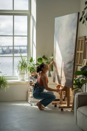 Focused artist woman looks down at picture studying details coming up with new ideas for creative. Interested pensive female painter adjusts canvas on easel to continue painting oil for exhibition.