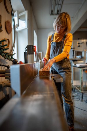 Careful focused carpenter woman works on electric planer wearing safety glasses. Concentrated female joiner grinds wooden block for handmade wooden furniture. Equipment for woodworking small business.