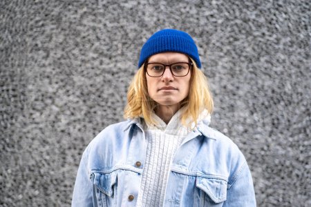 Photo for Long-haired blond hipster guy with stylish glasses standing in city street over grey wall, looking at camera. Closeup portrait of fashionable male looks like model, international student, tourist - Royalty Free Image