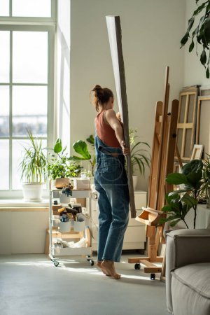 Interested female artist places canvas on easel to continue painting in oils in art studio. Focused woman in denim overalls barefoot holding, rearranges acrylic painting in craft studio on sunny day.