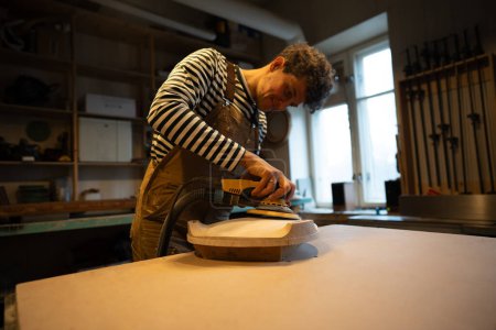 Sanding wood with orbital sander at workshop. Focused man carpenter polishes wooden seat of a future chair with electric sander. Carpentry workshop. Furniture production