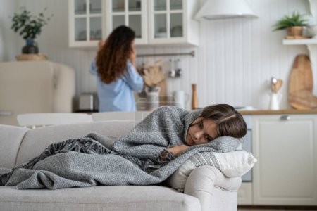 Sick unhappy schoolgirl wrapped in warm plaid lying on sofa with flu symptoms while her mother calling doctor by phone on background, kid with high fever at home. Children healthcare concept