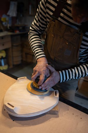 Nahaufnahme des Holzschleifens mit Orbitalschleifer in der Werkstatt. Tischler poliert Holzsitz eines zukünftigen Stuhls mit Elektroschleifer. Tischlerei. Möbelproduktion