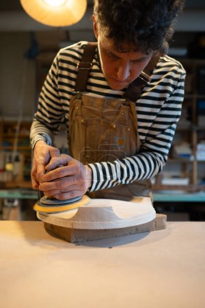 Lijado de madera con lijadora orbital en primer plano del taller. El carpintero masculino enfocado pule el asiento de madera de una futura silla con lijadora eléctrica. Taller de carpintería. Producción de muebles