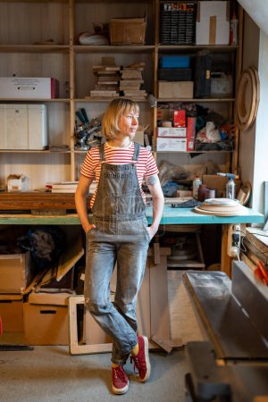 Serene woman carpenter take break after production wooden DIY furniture in joinery workshop standing with hands in pockets overalls. Relaxed crafts female resting thinking about new woodwork skills.