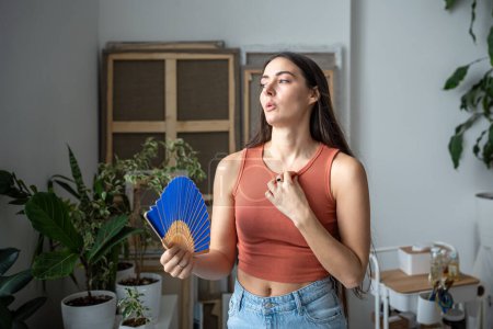 Overheated tired woman standing in room at hot summer weather day feels discomfort, suffers from heat waving blue hand fan to cool. Exhausted sweating dwelling female without air conditioner at home. 