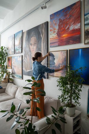 Photo for Woman artist hanging new painting on wall at loft studio. Girl assesses overall composition in apartment. Actual solutions by decoration in contemporary interior design. Artists cosy home. - Royalty Free Image