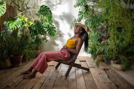 Carefree relaxed african american woman take break surround by tropical indoor plants. Satisfied young black female rest sits barefoot with closed eyes on chair in urban jungle home with houseplants.