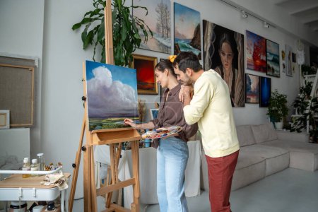 Creative woman painter drawing picture in art studio, husband enjoys process, good relationship. Loving man support wife female artist paints landscape canvas on easel standing at home craft workshop.