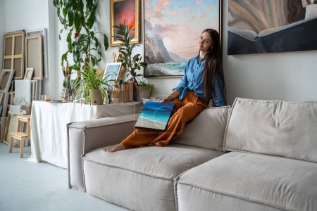 Photo for Creative pleased woman artist demonstrates new painted picture for gallery, sitting on couch at art studio. Relaxed joyful female painter sharing painting feeling satisfaction result at home workshop - Royalty Free Image