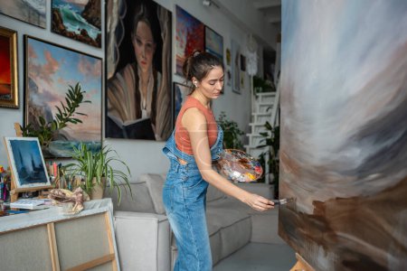 Konzentriert inspirierte Künstlerin zeichnet auf Leinwand im Kunstatelier mit Zimmerpflanzen. Die interessierte junge Malerin mit Farbpalette und Pinsel trägt Ölfarbe auf die komplette Malerei auf, auf Staffelei.
