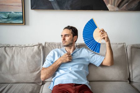 Téléchargez les photos : Triste homme se ventilant avec ventilateur bleu dans l'appartement. Épuisé souffrant de chaleur, assis sur le canapé, pas de climatiseur à la maison. Température chaude, mauvais temps d'été, concept de contrôle climatique - en image libre de droit
