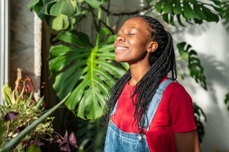 Téléchargez les photos : Femme noire positive profitant de rayons de soleil chaud parmi les plantes vertes. afro-américaine fille dans optimiste heureux humeur souriant largement et atteindre pour matin soleil sentiment d'énergie parmi la jungle urbaine - en image libre de droit