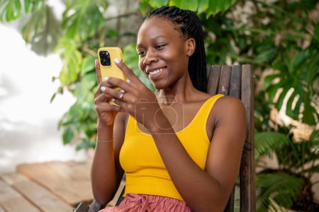 Téléchargez les photos : Fille africaine joyeuse avec smartphone. Souriant joyeuse femme afro-américaine noire utilisant le téléphone reposant parmi les feuilles de plantes vertes. Femme blogueur écrit des billets de blog, retourne à travers le flux sur les réseaux sociaux - en image libre de droit