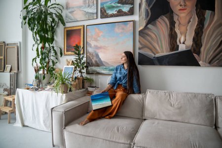 Dreamy female artist sitting on back of sofa, holding painting in hand, looking out window. Woman takes break from creative work, seeking for inspiration, relaxing resting at home art studio.