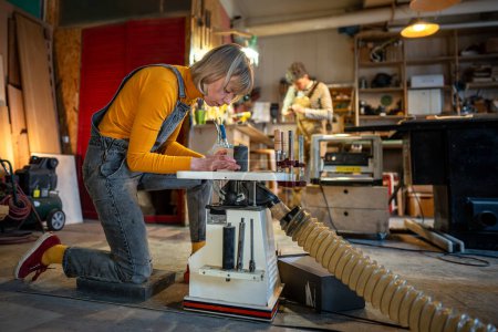 Photo for Concentrated carpenter woman carefully turns wooden products on lathe. Craftswoman works in family joinery workshop polishing woodworking items with special machine for handmade furniture for order. - Royalty Free Image
