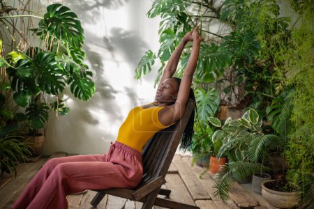 Sleepy african american young woman stretching out on chair surround by tropical houseplants. Relaxed tired black girl gardener puts hands up resting on wooden armchair after work with indoor plants.