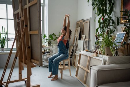 Entspannte Künstlerin auf Stuhl sitzend und tief seufzend aus Fenster schauend. Ruhige, kraftvolle, enthusiastische Frau lässt sich inspirieren, bevor sie mit dem produktiven Arbeitstag an einem großen Kunstprojekt beginnt.