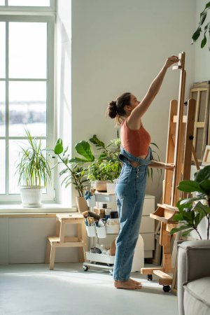 Creative female artist adjusting height of easel before starting work. Inspired woman preparing workplace workspace for oil painting process on canvas. Girl painter in sunny apartment home art studio.