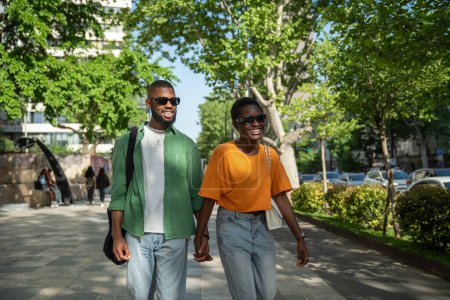 Cheerful carefree african american couple walks with hold hands in sidewalk downtown guy makes girlfriend laugh by fun storytelling. Stylish serene black young woman enjoy stroll with happy boyfriend.