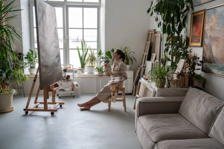 Perfectionist woman artist critically evaluates own painting, sitting on chair, looking for flaws, minuses in work done. Serious strict female painter thinks carefully to refine picture to ideality.