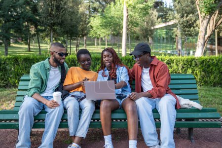 Photo for Contented group african american students of casually seated on park bench, girls balances laptop on knees looks at screen with guys. Interested black friends prepare for exam, test together outdoors. - Royalty Free Image