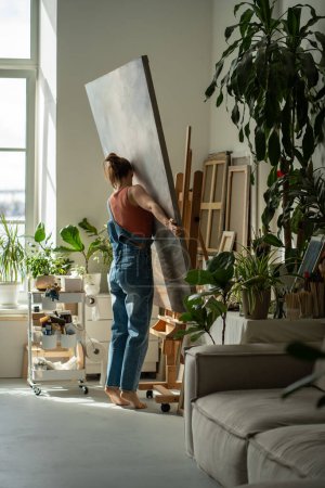Female artist carries, places canvas on easel to continue painting in oils in art studio. Focused woman in denim overalls barefoot holding, rearranges acrylic painting in craft studio on sunny day.