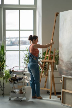 Focused female artist adjusting height of easel before starting work. Inspired woman preparing workplace workspace for oil painting process on canvas. Girl painter in sunny apartment home art studio.