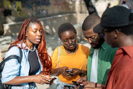 Interesado cuatro estudiantes afroamericanos niñas y chicos sosteniendo teléfonos inteligentes que comparten información de estudio de pie al aire libre juntos. Grupo serio enfocado de amigos negros mira en el teléfono de la pantalla fuera.